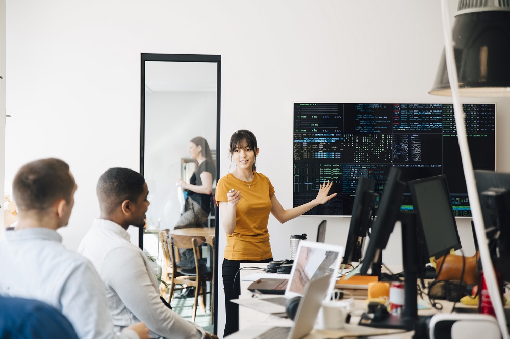 Eine ITlerin ist mit zwei Kollegen in einem hellen, modernen Büro. Sie steht vorne an einem Bildschirm, auf dem Code zu sehen ist und erklärt etwas.