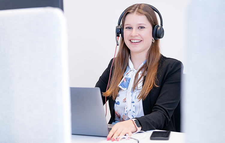 Mitarbeiterin sitzt mit Headset lächelnd vor ihrem Laptop am Schreibtisch